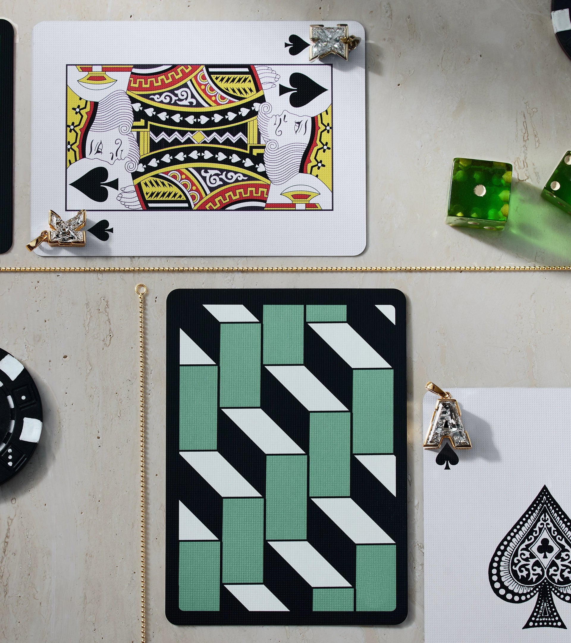 High-angle lifestyle shot of ALTR Created Diamond letter pendants shaped like 'A' and 'K' on playing cards, featuring a King of Spades, green dice, and a poker chip on a stone surface.