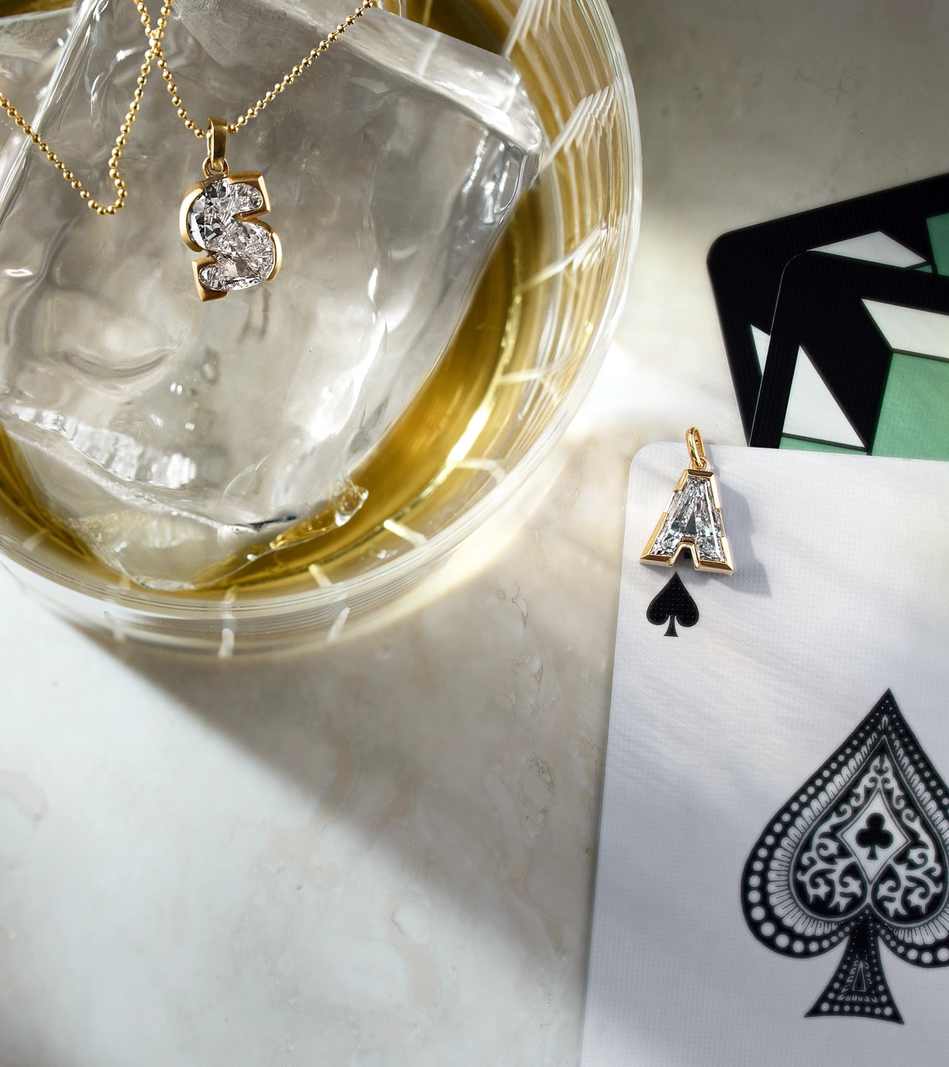 Luxury lifestyle photography of ALTR lab-grown diamond initial pendants 'S' and 'A' in yellow gold settings, styled next to a cocktail glass with ice and an Ace of Spades playing card.