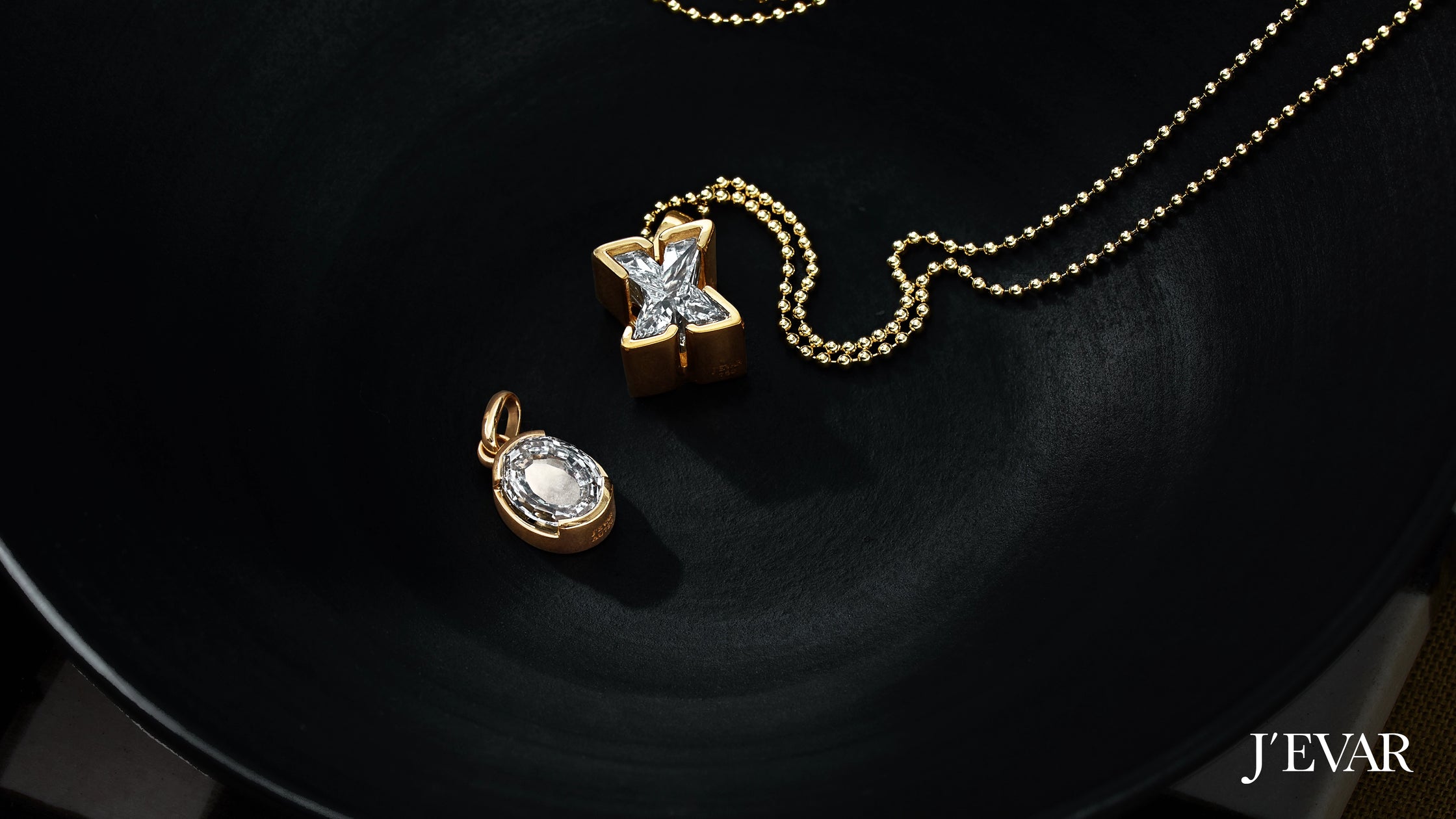 Close-up of gold Jevar's TYPE solitaire initial diamond letter pendants in 'X' and 'O' set on a textured black plate.