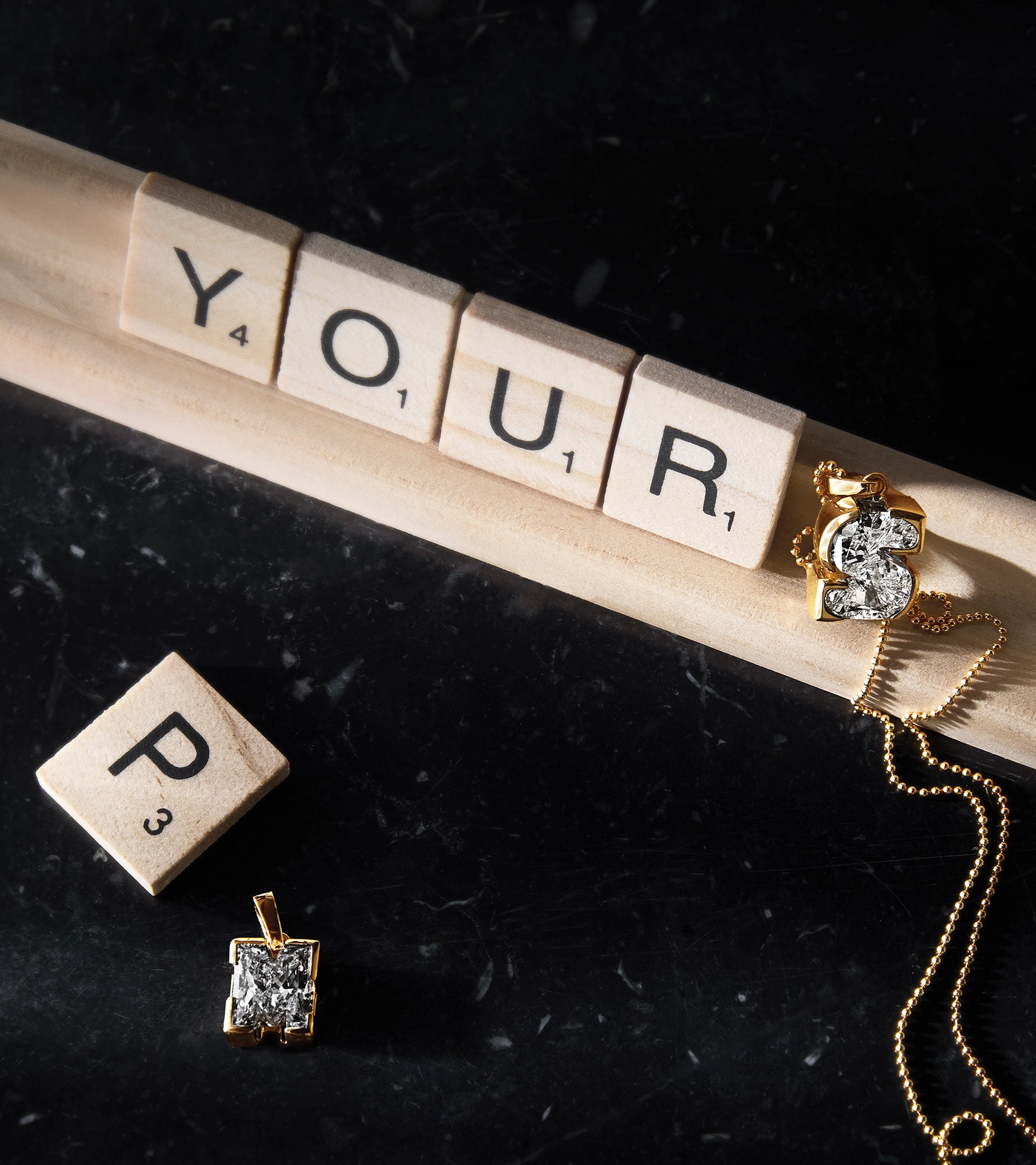 Wooden letter tiles in a game of Scrabble spelling "YOUR" next to a Jevar TYPE 'S' solitaire initial diamond letter pendant in 18k recycled gold on a dark marble surface.