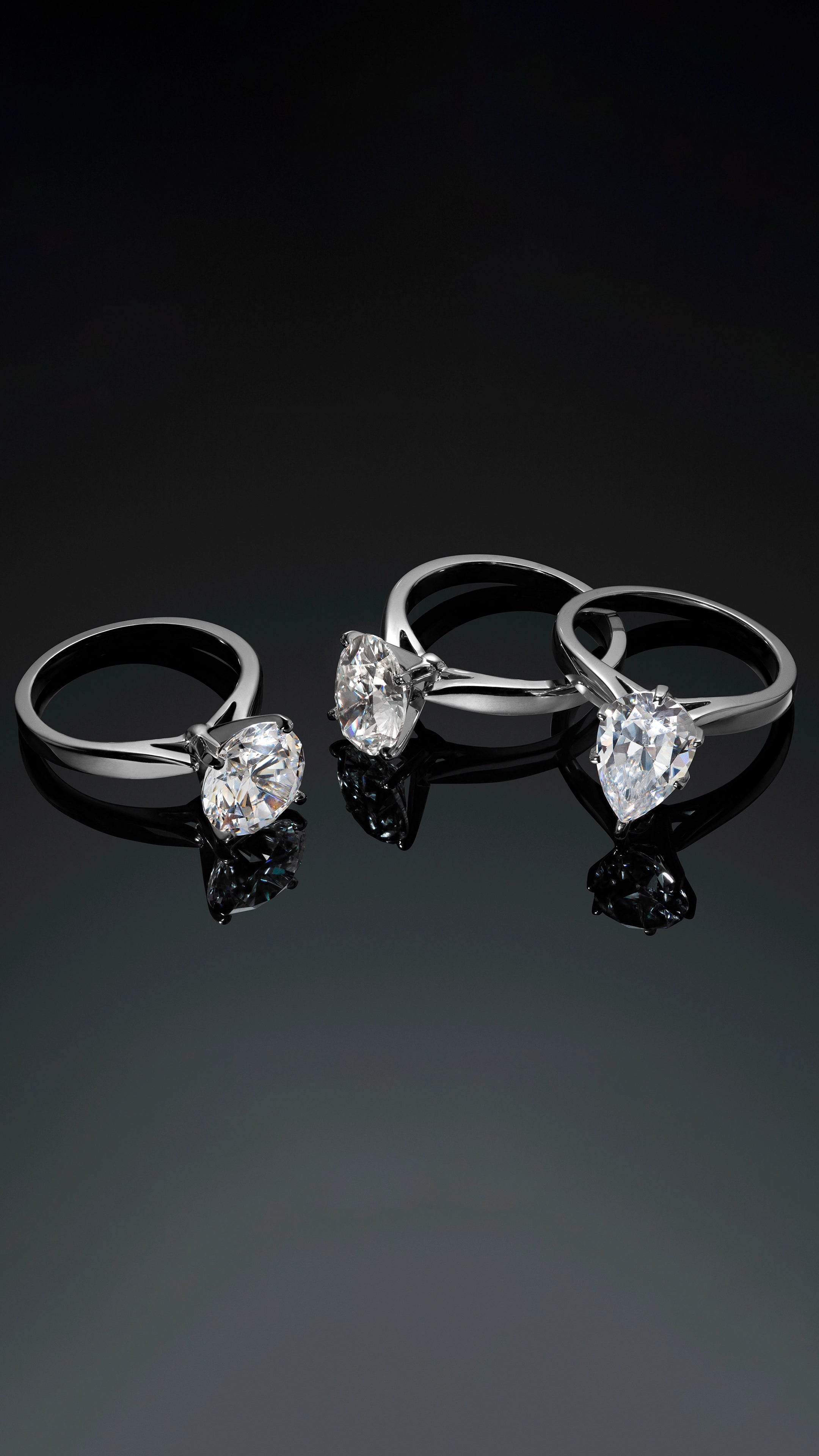 Three round and pear-cut lab-grown diamond solitaire rings on a reflective black surface.