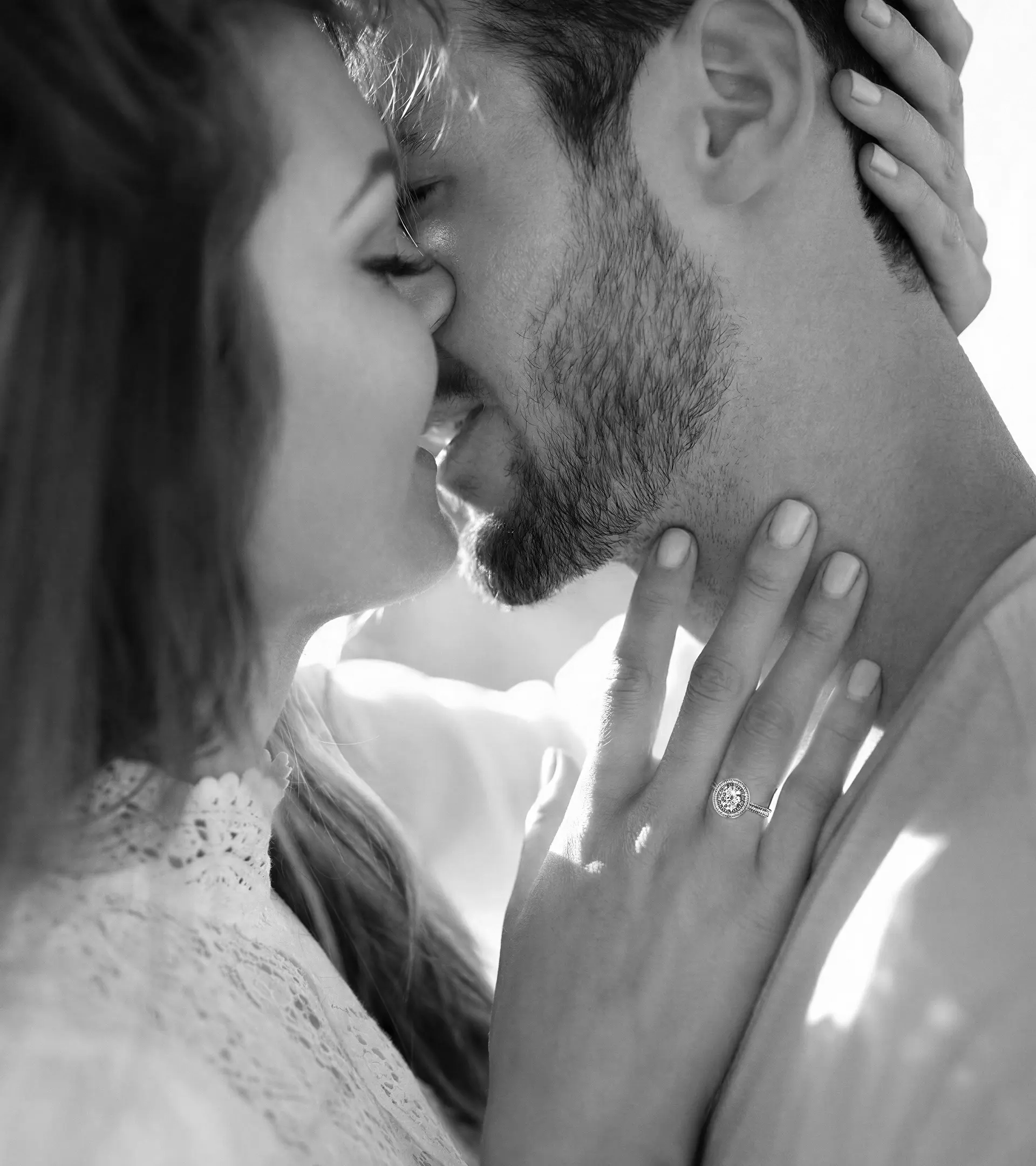 Black and white lifestyle shot of a romantic moment, featuring a woman’s hand with a round-cut halo diamond engagement ring resting on a man’s neck.